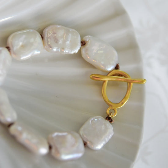 Close-up of a bracelet with irregular white pearls linked by brown cord, featuring a gold toggle clasp, set on a white textured background. Luxurious and elegant.