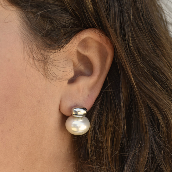 Close-up of an ear wearing a large, elegant pearl earring with a silver top. The earring contrasts against the skin and dark brown hair.