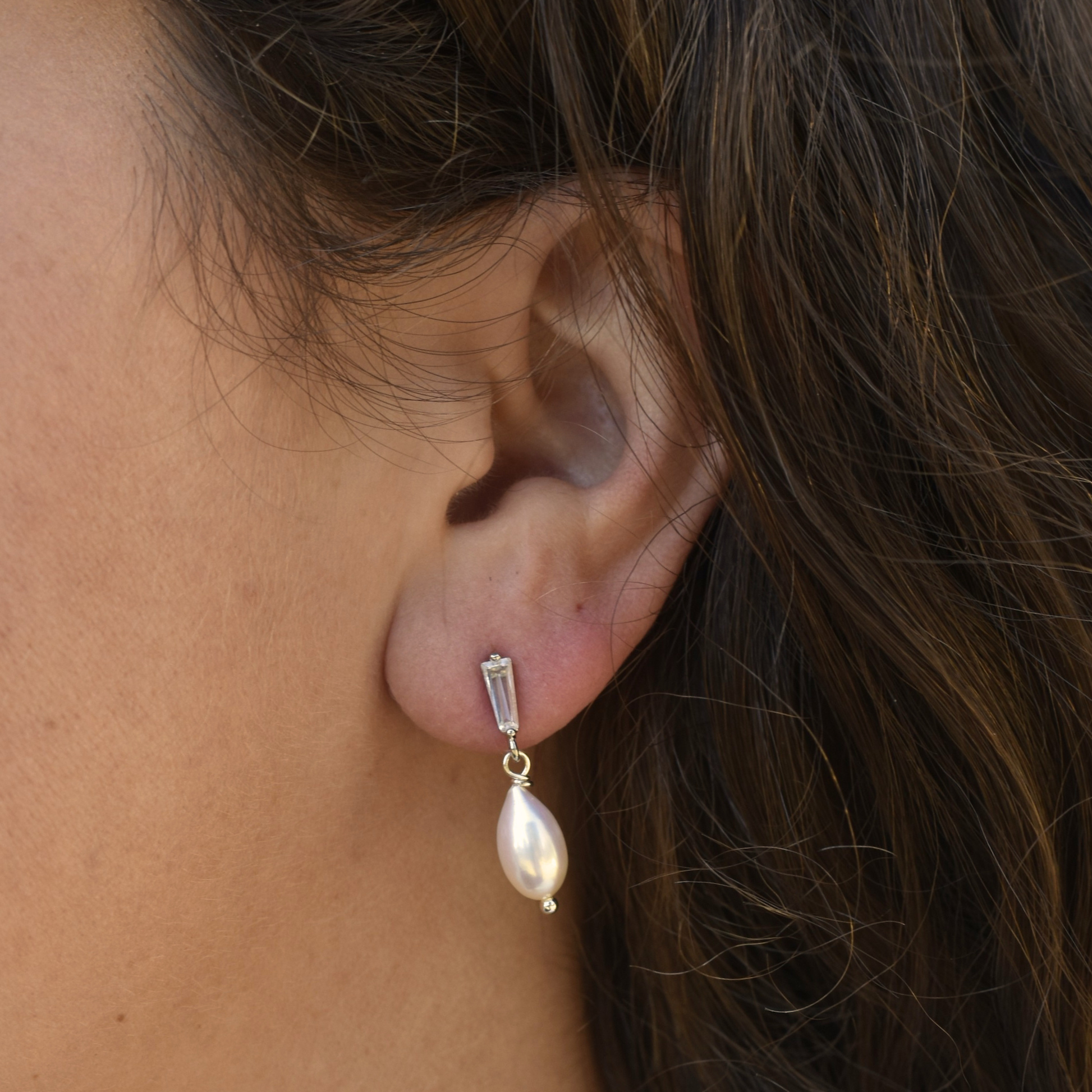 Close-up of an ear wearing a large, elegant pearl earring with a gold top. The earring contrasts against the skin and dark brown hair.