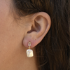 Close-up of an ear wearing an elegant earring. The earring features a rectangular gemstone and a dangling, irregularly shaped pearl, set in gold.
