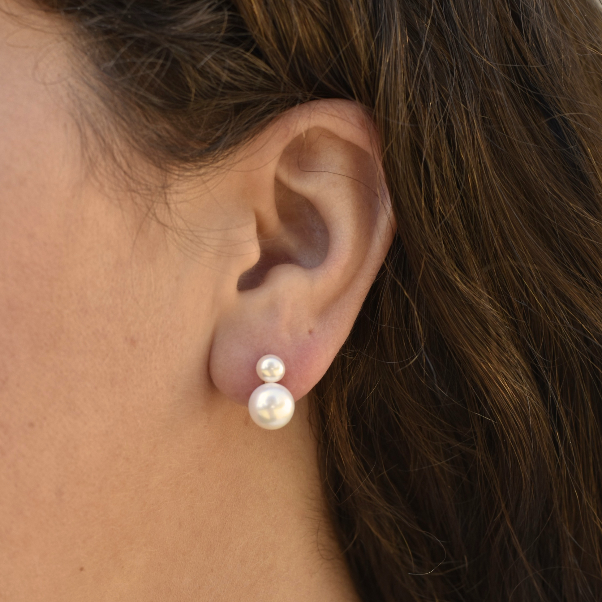 Close-up of an ear wearing elegant double pearl earrings. The larger pearl dangles below a smaller one, on a backdrop of brown hair.