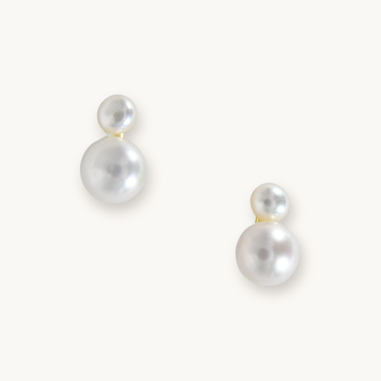 Elegant pearl earrings with two round, white pearls of varying sizes on each earring. They rest on a light background, conveying simplicity and sophistication.