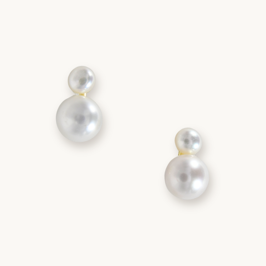 Elegant pearl earrings with two round, white pearls of varying sizes on each earring. They rest on a light background, conveying simplicity and sophistication.