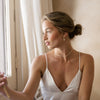 Woman in a white satin slip sitting by a window with light curtains. Dainty wedding jewelry for bridal look. 