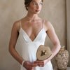 Woman in a white dress gazes to the side, hair in an elegant bun, and she's holding a small vintage mirror. She's wearing flower shaped pearl earrings and a simple choker, with a serene expression.