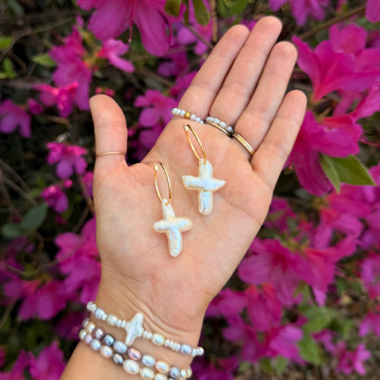 A hand displays cross-shaped pearl earrings against vibrant pink flowers. The wrist is adorned with pearl bracelets, conveying elegance and a touch of nature.
