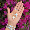 A hand displays cross-shaped pearl earrings against vibrant pink flowers. The wrist is adorned with pearl bracelets, conveying elegance and a touch of nature.