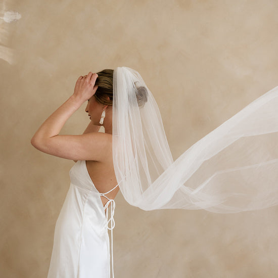 Bride in a white dress and flowing veil, adjusting her hair against a neutral background. The scene conveys elegance and serenity.
