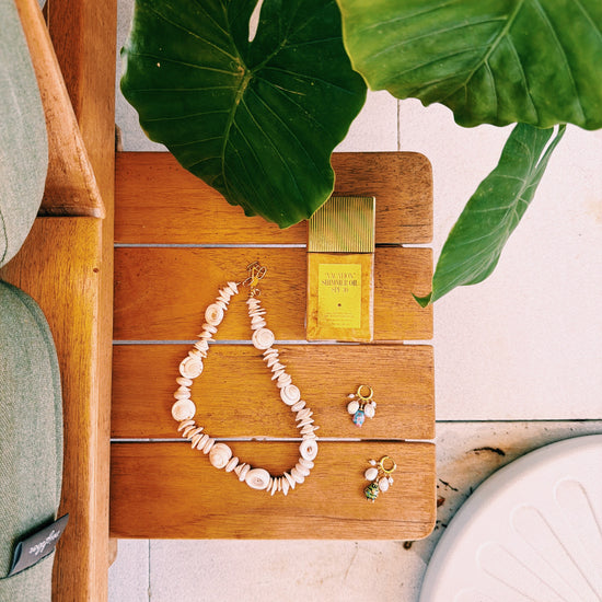 A flat lay of a shell necklace, two pairs of pearl earrings, and a gold bottle labeled "SHIMMER OIL" on a wooden surface. Large green leaves add a natural touch.