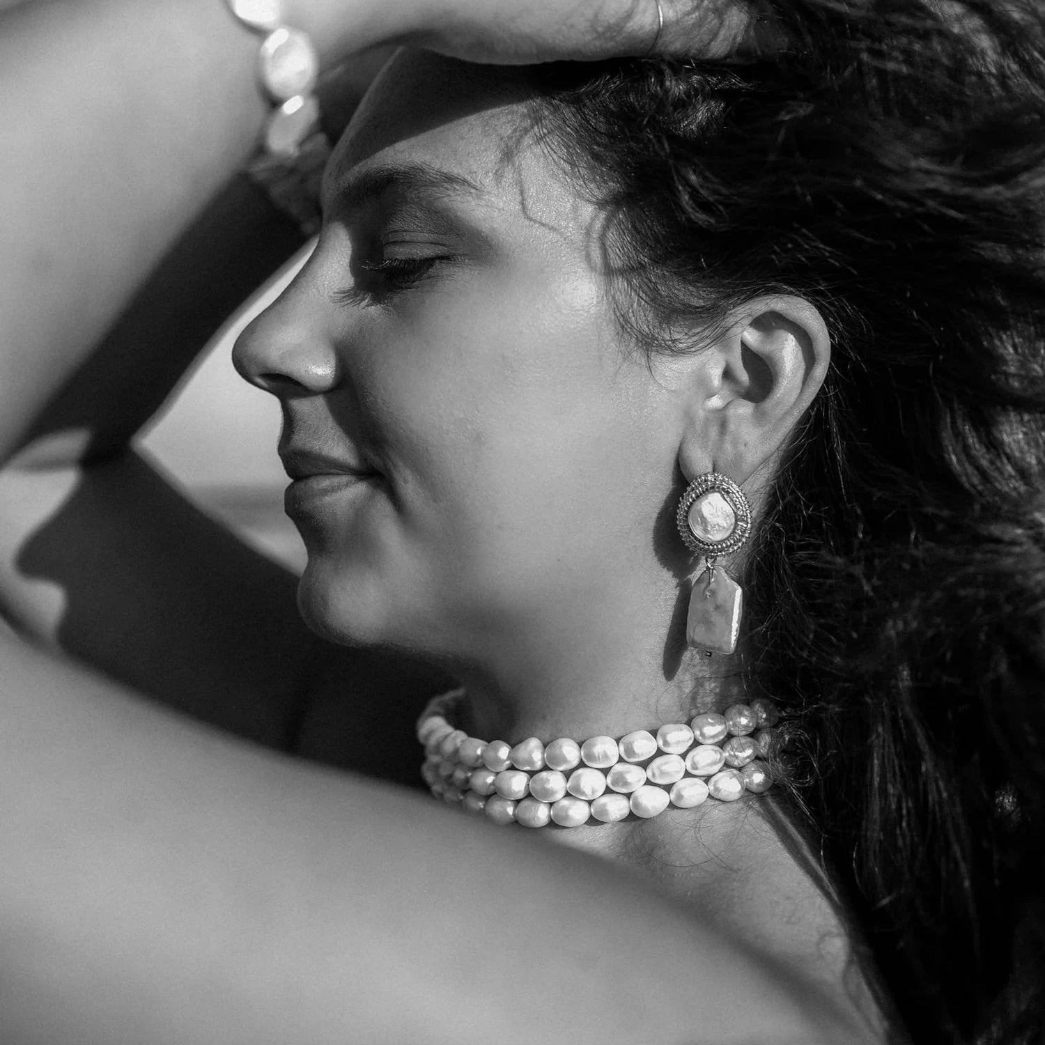 Black and white portrait of a woman in profile, eyes closed, expressing calm. She wears pearl jewelry, including earrings and a choker.