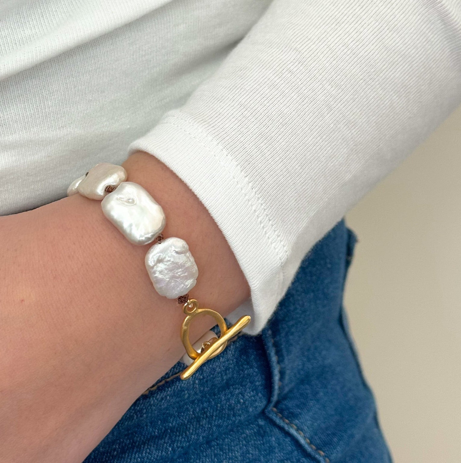 A close-up of a wrist wearing a white long-sleeve shirt and blue jeans. The wrist features a bracelet with irregular white pearls and a gold toggle clasp.