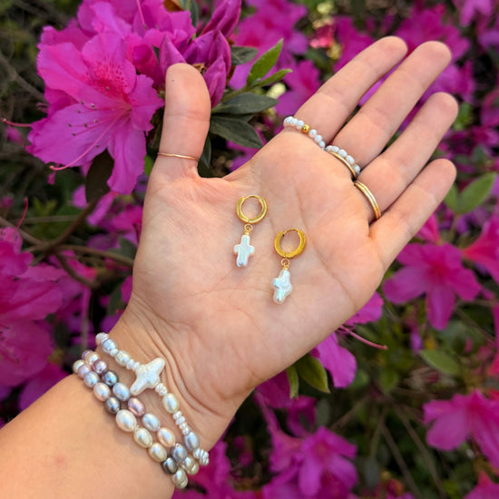 A hand displays cross-shaped pearl earrings against vibrant pink flowers. The wrist is adorned with pearl bracelets, conveying elegance and a touch of nature.