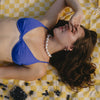 Woman in blue bikini top and pearl necklace lies on yellow checkered blanket, covering eyes, beside scattered blueberries on a sandy beach.