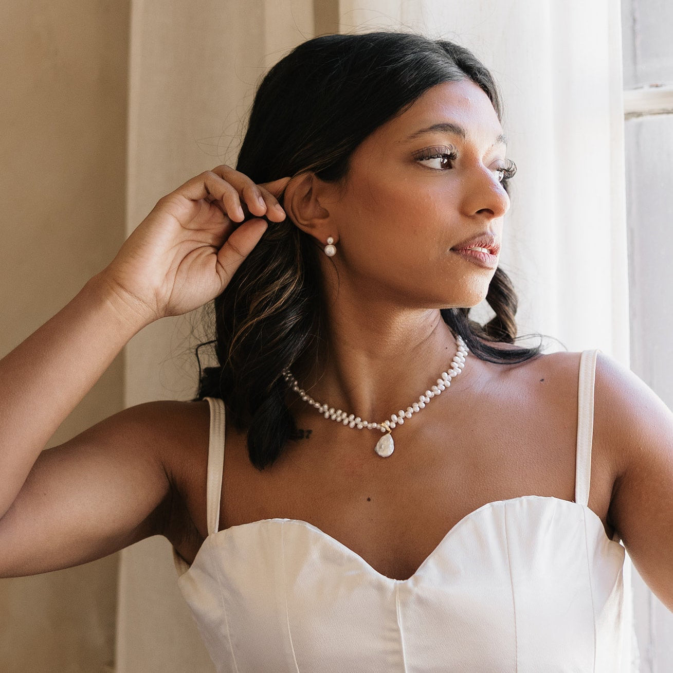Woman wearing  timeless bridal pearl necklace and earrings, adjusting her hair in a softly lit room.