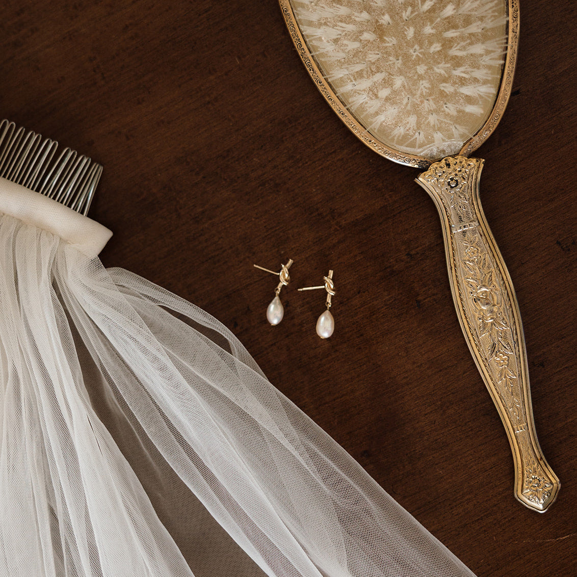 Ornate vintage hair brush and bridal veil comb on dark wood. Gold knots with tear-shaped pearl earrings add elegance to the nostalgic, romantic scene.