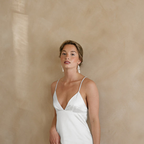 Young woman in a white satin dress with thin straps, wearing statement tassel earrings. She stands against a soft beige background, exuding elegance.