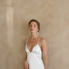 Young woman in a white satin dress with thin straps, wearing statement tassel earrings. She stands against a soft beige background, exuding elegance.