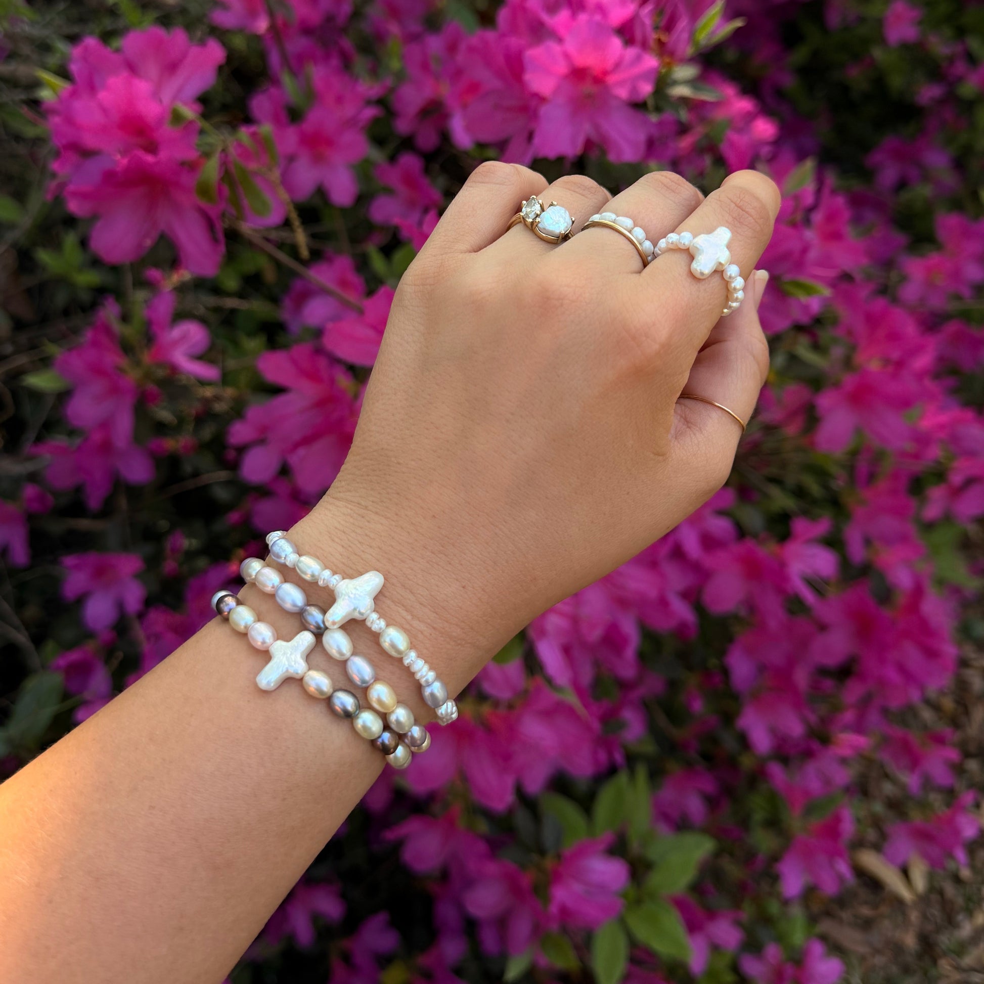 A hand adorned with pearl bracelets and rings is held against vibrant pink flowers. The jewelry exudes elegance and the scene feels tranquil.