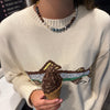 A person wearing a cream knit sweater with a brown and green graphic design on the chest, holding an ice cream cone in a waffle cone. The sweater features a small embroidered logo on the lower right of the graphic.