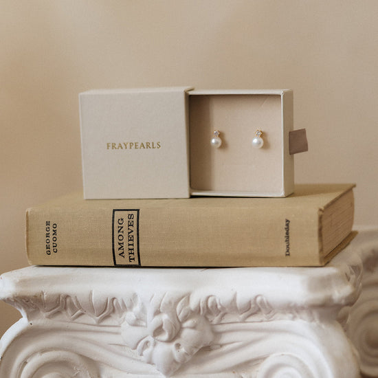 Pair of earrings in a box on top of a book titled 'Amour et Maitrise' on a decorative white pedestal.