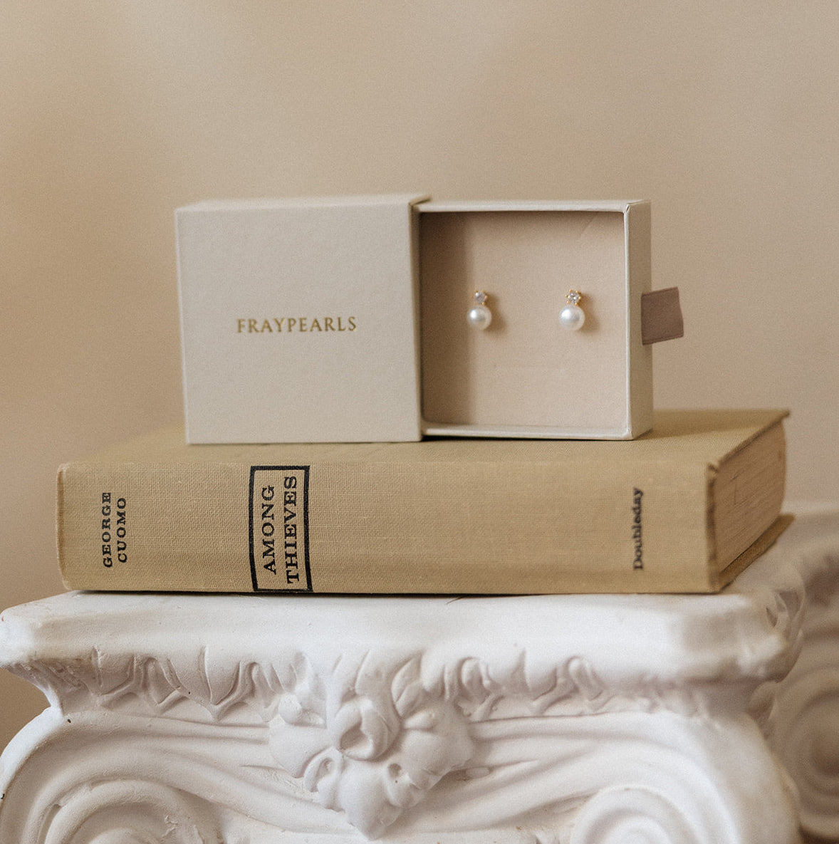 Pair of earrings in a box on top of a book titled 'Amour et Maitrise' on a decorative white pedestal.