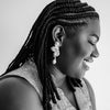 Black and white close-up of a person smiling, wearing braided hair and a large, intricate earring shaped like a flower. The mood is joyful and serene.