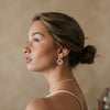 Profile of a woman with a contemplative expression, wearing pearl earrings and a necklace. Her hair is styled in a low bun against a soft beige background.