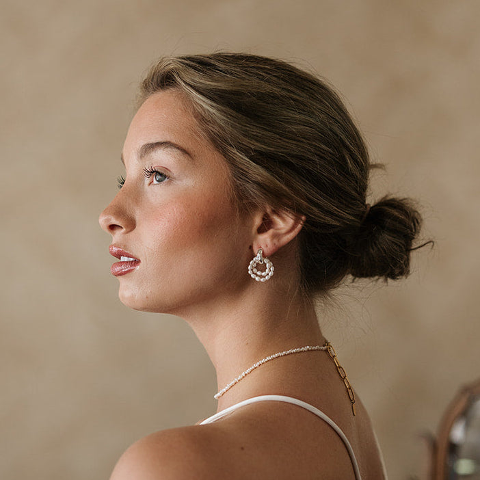 Profile of a woman with a contemplative expression, wearing pearl earrings and a necklace. Her hair is styled in a low bun against a soft beige background.