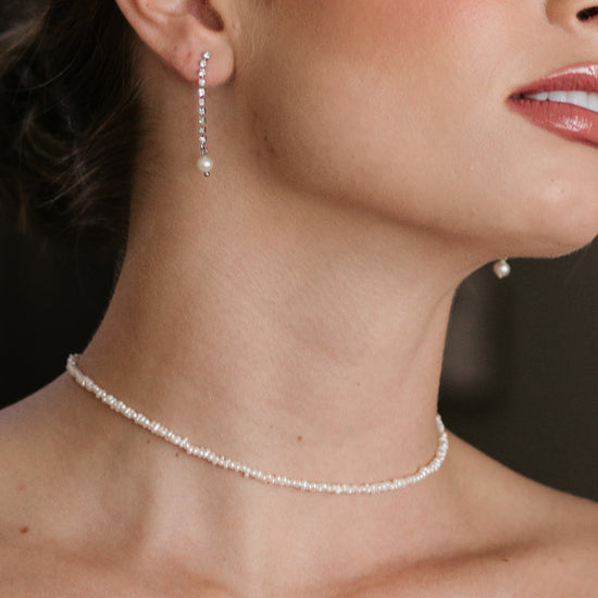 Close-up of a woman's neck and jawline wearing delicate pearl earrings and a matching pearl necklace. Skin appears smooth, with a soft, elegant tone.