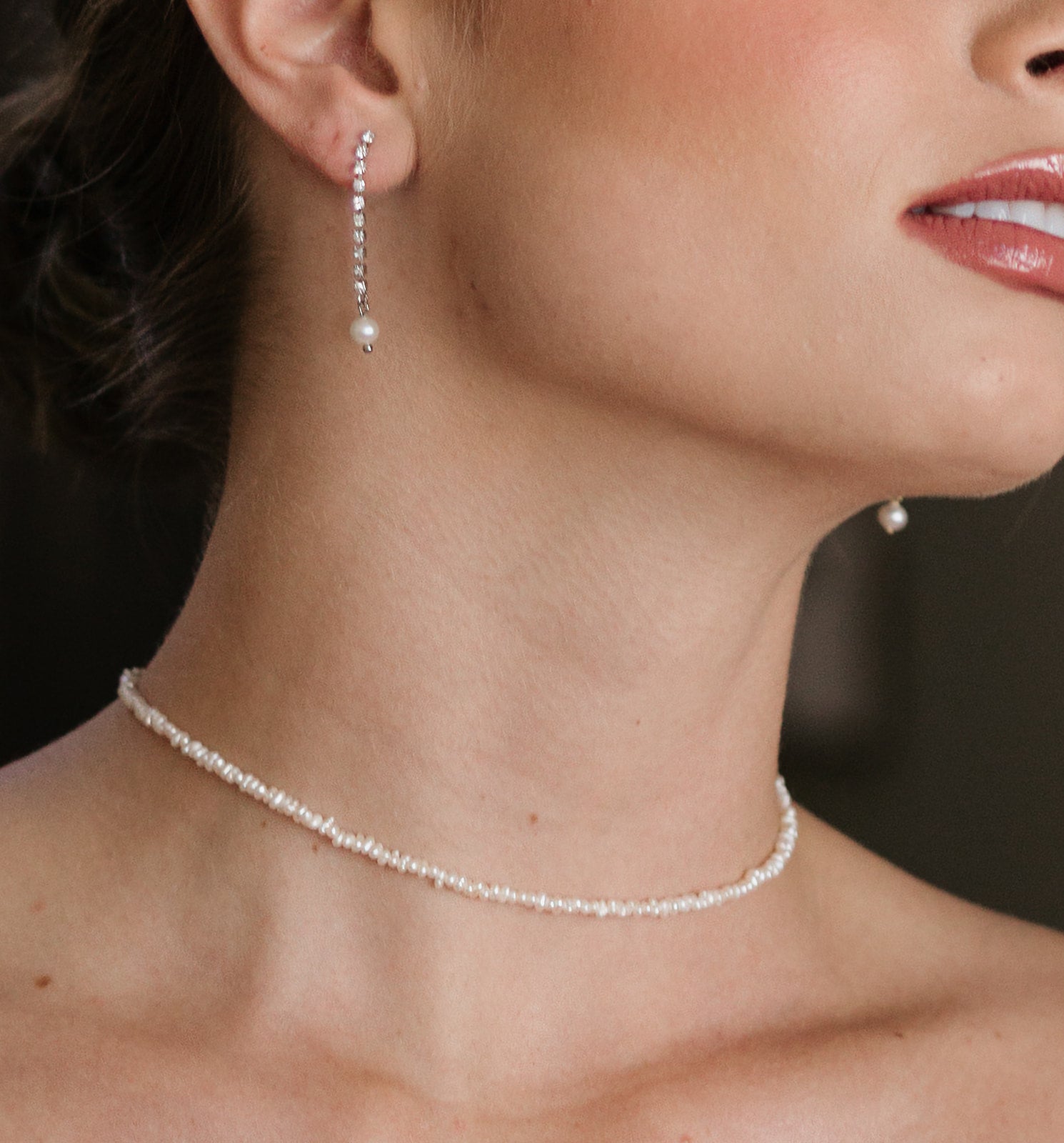 Close-up of a woman's neck and jawline wearing delicate pearl earrings and a matching pearl necklace. Skin appears smooth, with a soft, elegant tone.
