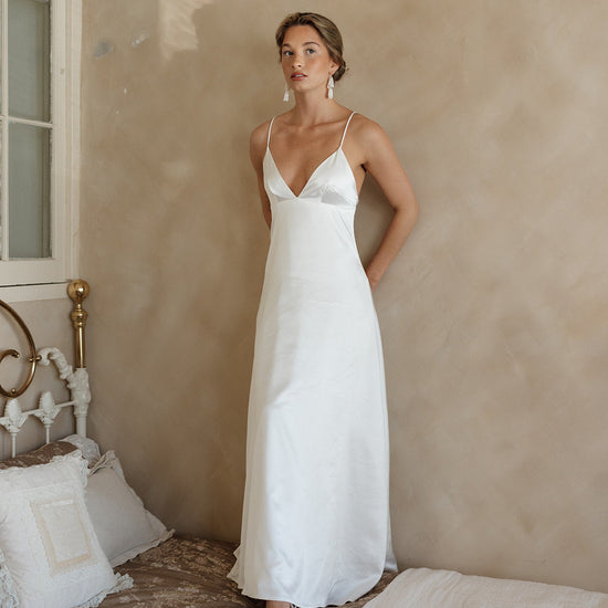 Woman wearing a white satin gown with thin straps, standing against a beige wall. She has a serene expression, exuding an elegant and calm vibe.