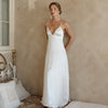 Woman wearing a white satin gown with thin straps, standing against a beige wall. She has a serene expression, exuding an elegant and calm vibe.
