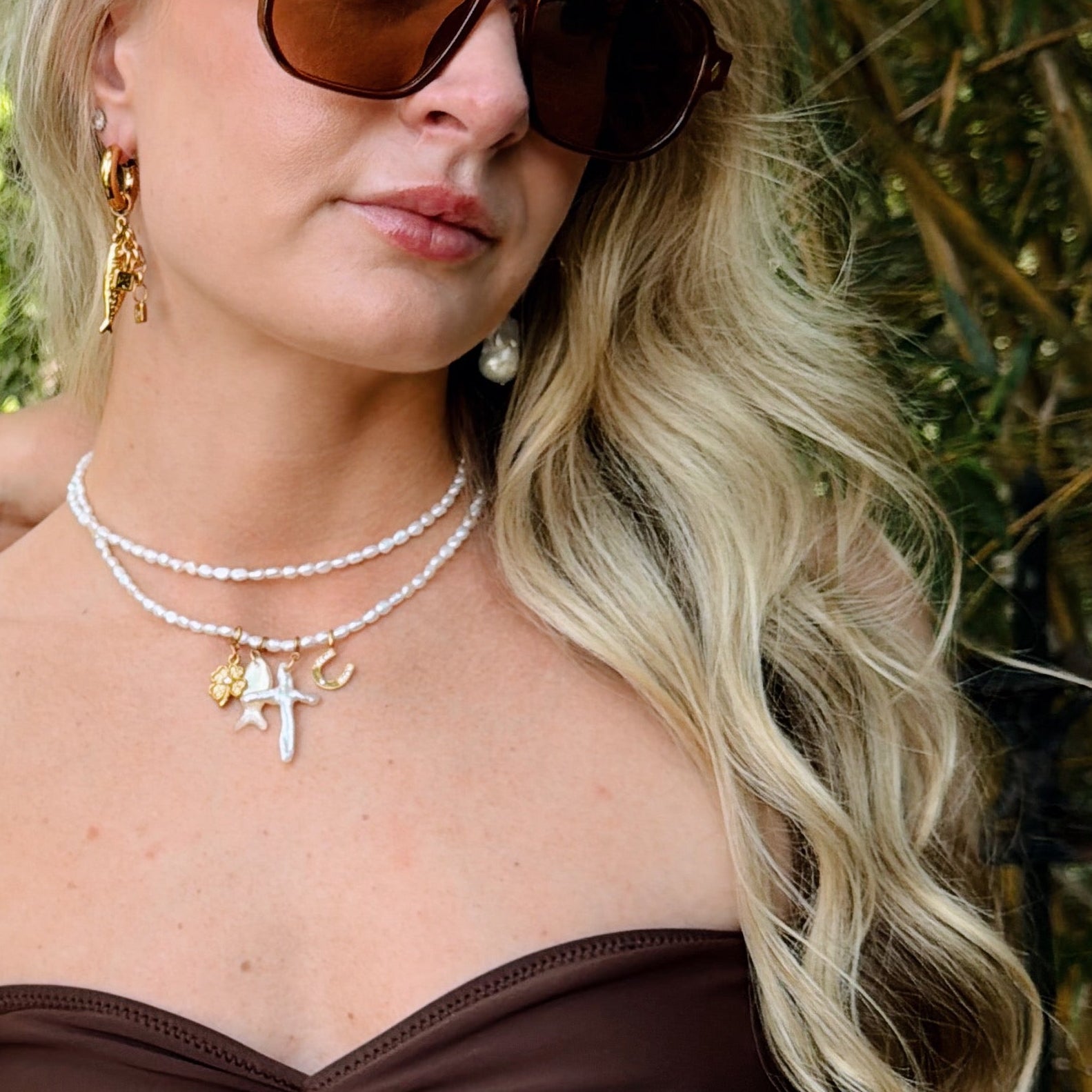 A woman with blonde hair wears large brown sunglasses, a brown strapless top, and layered pearl necklaces with charms. She has gold and pearl earrings.