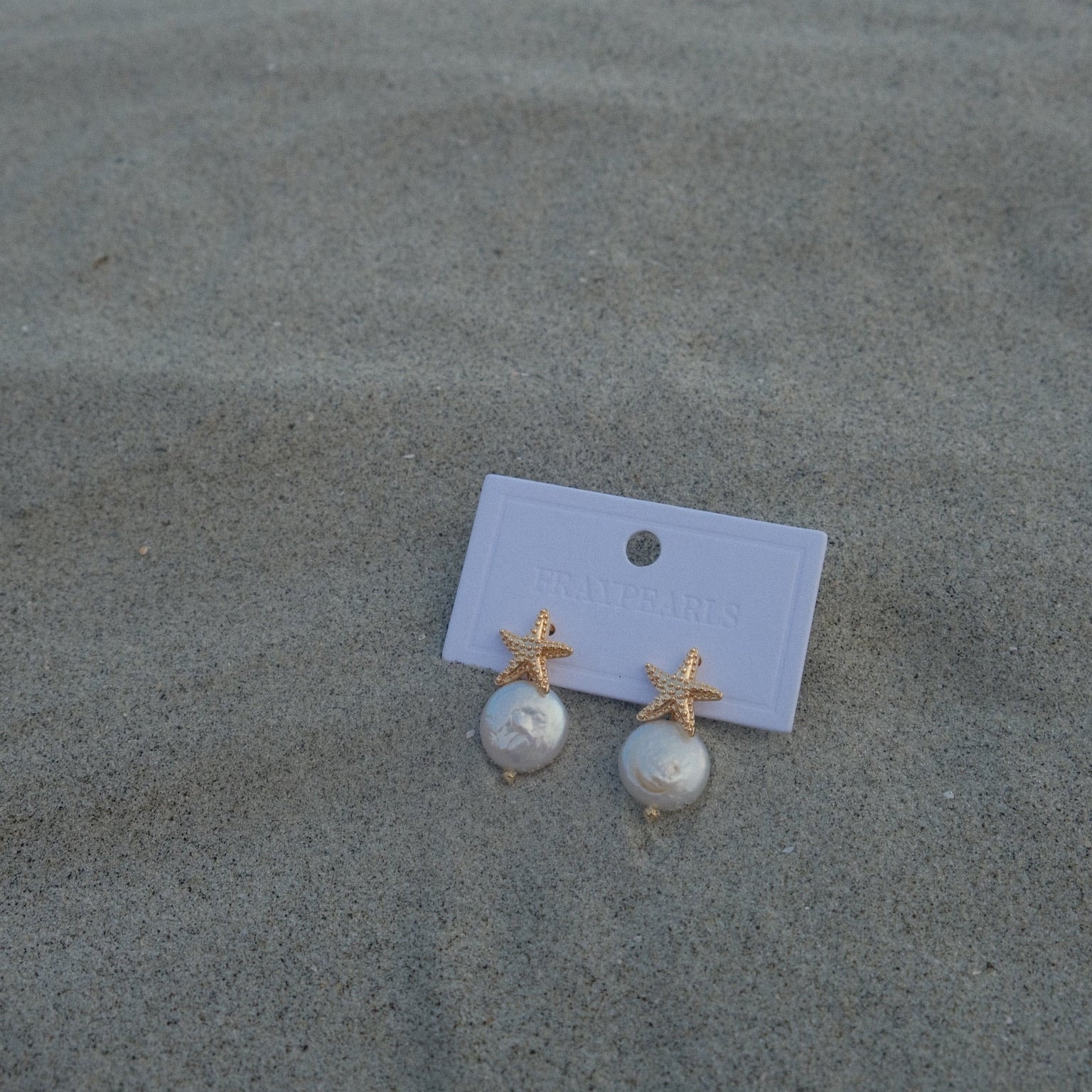 Elegant earrings with gold starfish and round pearls on a white card, displayed on textured sand, evoke a seaside charm.