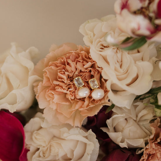 Elegant pearl and gold earrings rest on a peach carnation, surrounded by white and red roses, creating a soft, romantic atmosphere.