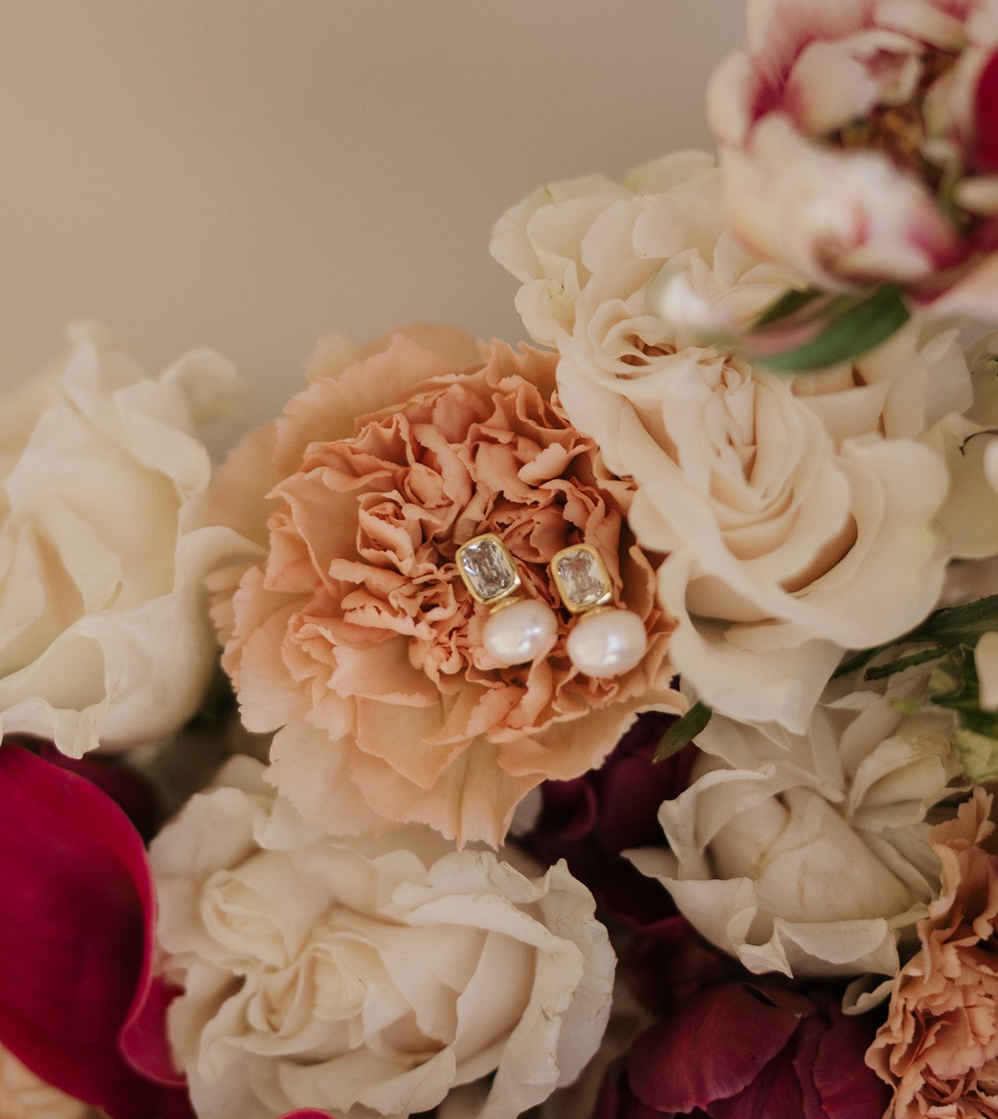 Elegant pearl and gold earrings rest on a peach carnation, surrounded by white and red roses, creating a soft, romantic atmosphere.