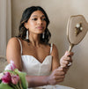 A woman in a white dress and pearl tassel earrings gazes into an ornate hand mirror with a thoughtful expression; soft lighting and pink flowers add elegance.