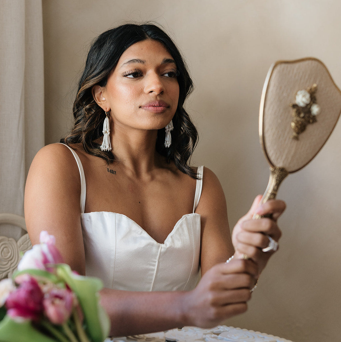 A woman in a white dress and pearl tassel earrings gazes into an ornate hand mirror with a thoughtful expression; soft lighting and pink flowers add elegance.