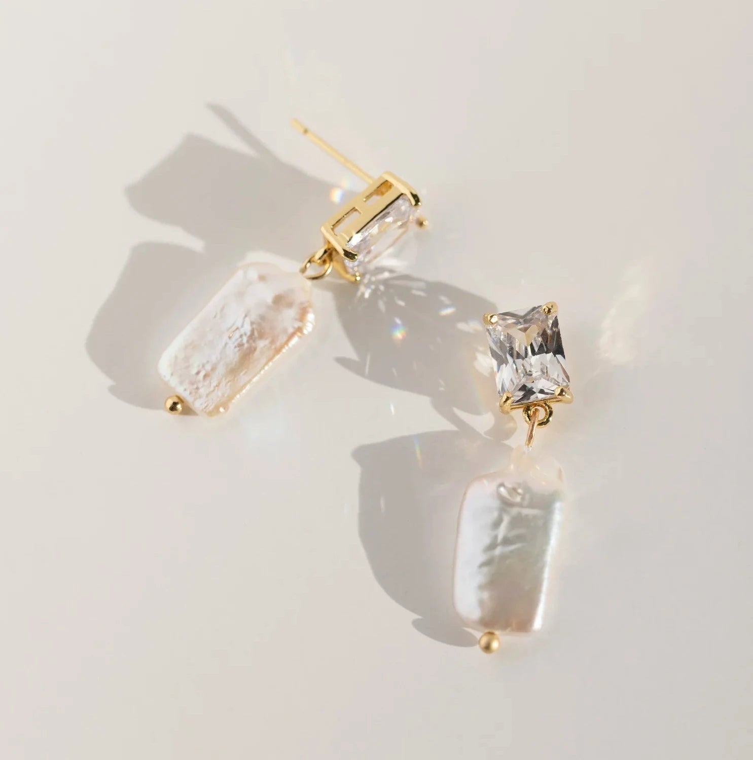 Elegant earrings with rectangular clear gemstones and iridescent pearl-like dangles set in gold, casting soft shadows on a neutral surface.