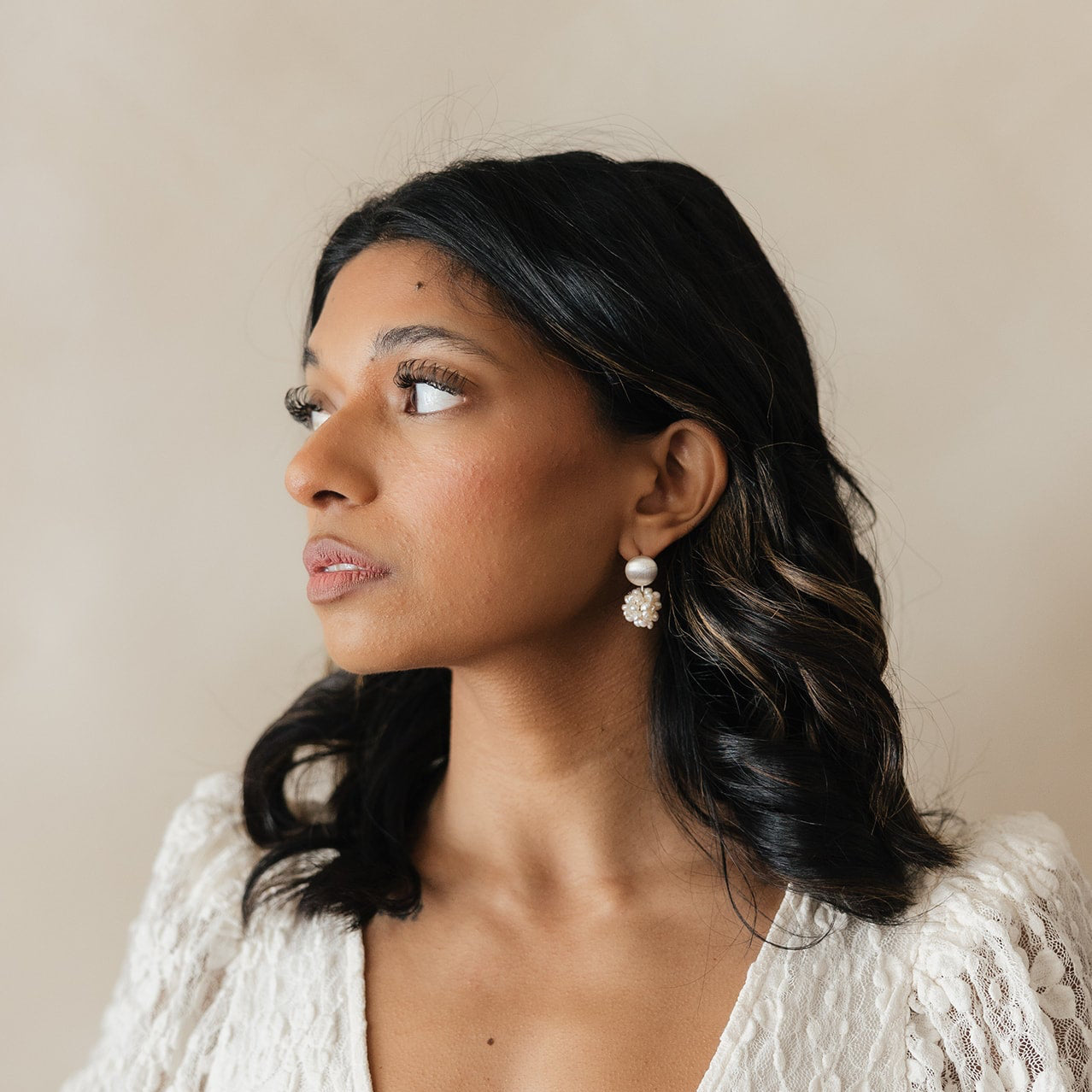 Woman in profile with wavy black hair and pearl earrings, wearing a white lace top. She gazes thoughtfully to the side against a soft beige background.