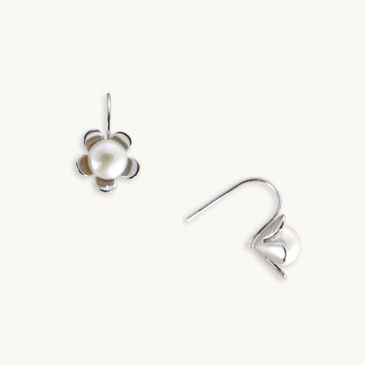 Silver earrings shaped like flowers, each with a central pearl. One earring is shown face-up, the other side view, on a plain, light background.