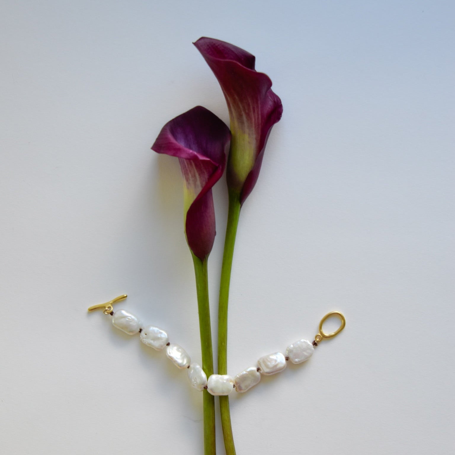 Two deep purple calla lilies with green stems are positioned against a white background. A pearl bracelet with gold clasps is draped around the stems.