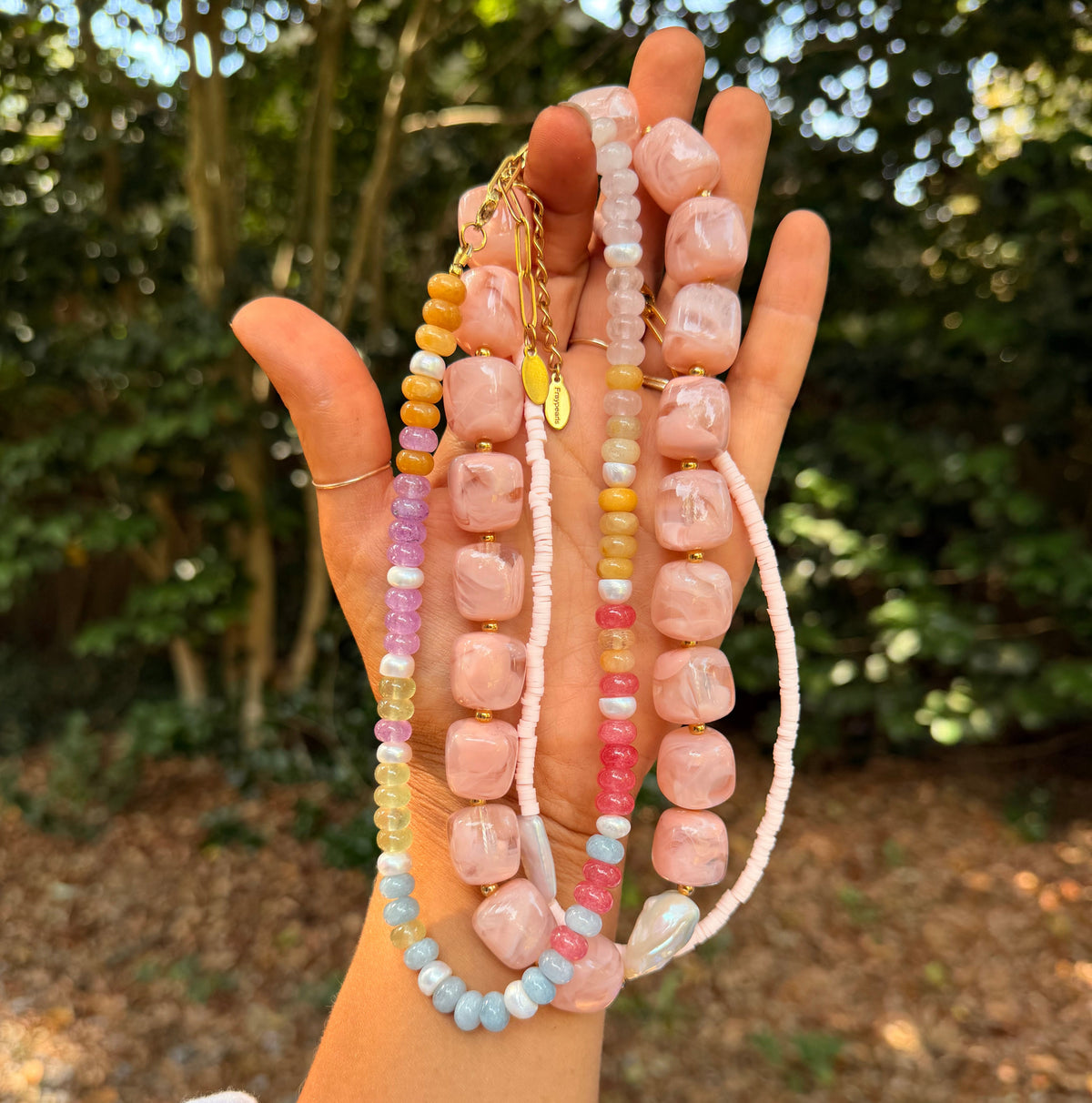 A hand holding three beaded necklaces outdoors, with a blurred background of trees and sunlight. The necklaces feature chunky pale pink stone beads, pastel multicolor beads, and a small gold charm.