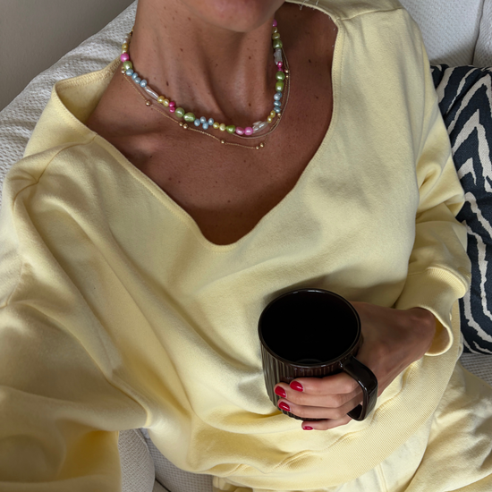 A person in a cozy yellow sweater holds a dark mug, displaying red-painted nails. They're wearing a colorful pearl necklace, evoking a relaxed and cheerful vibe.