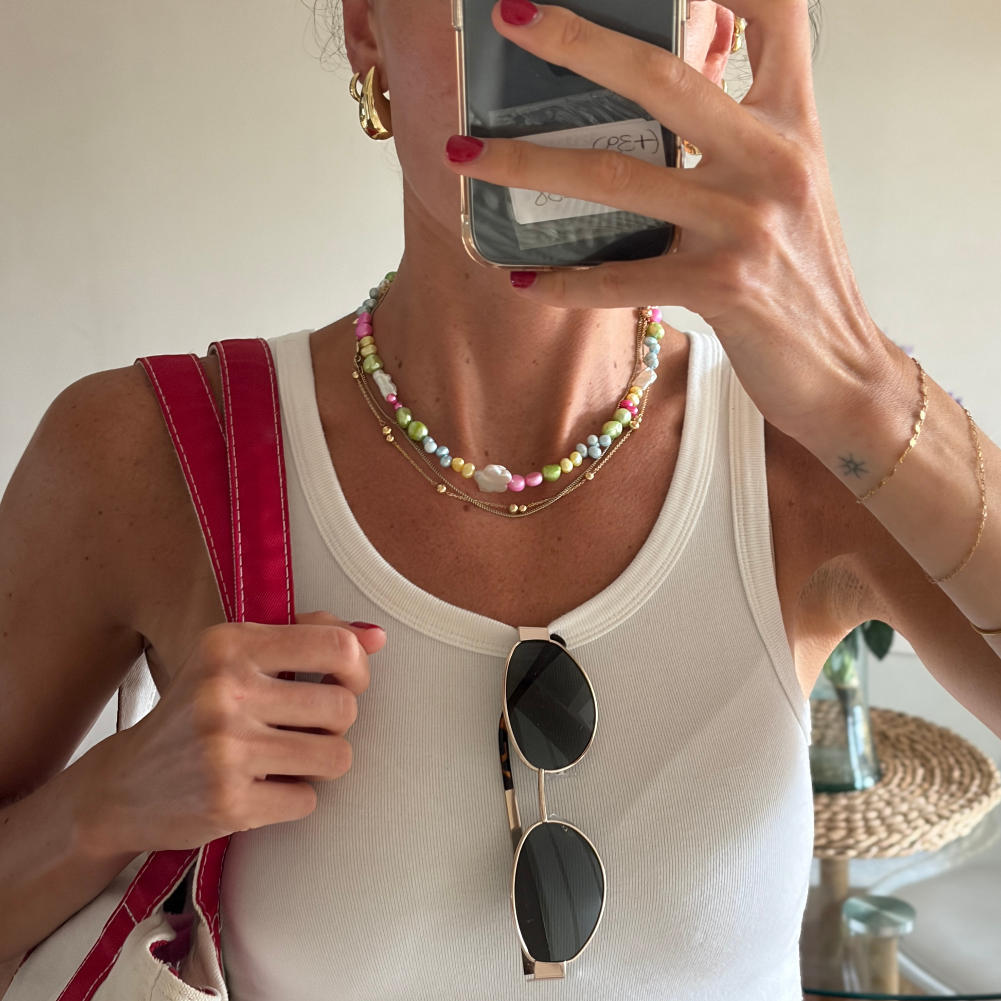 Person taking a mirror selfie wearing a white tank top, colorful pearl necklace, and holding sunglasses.