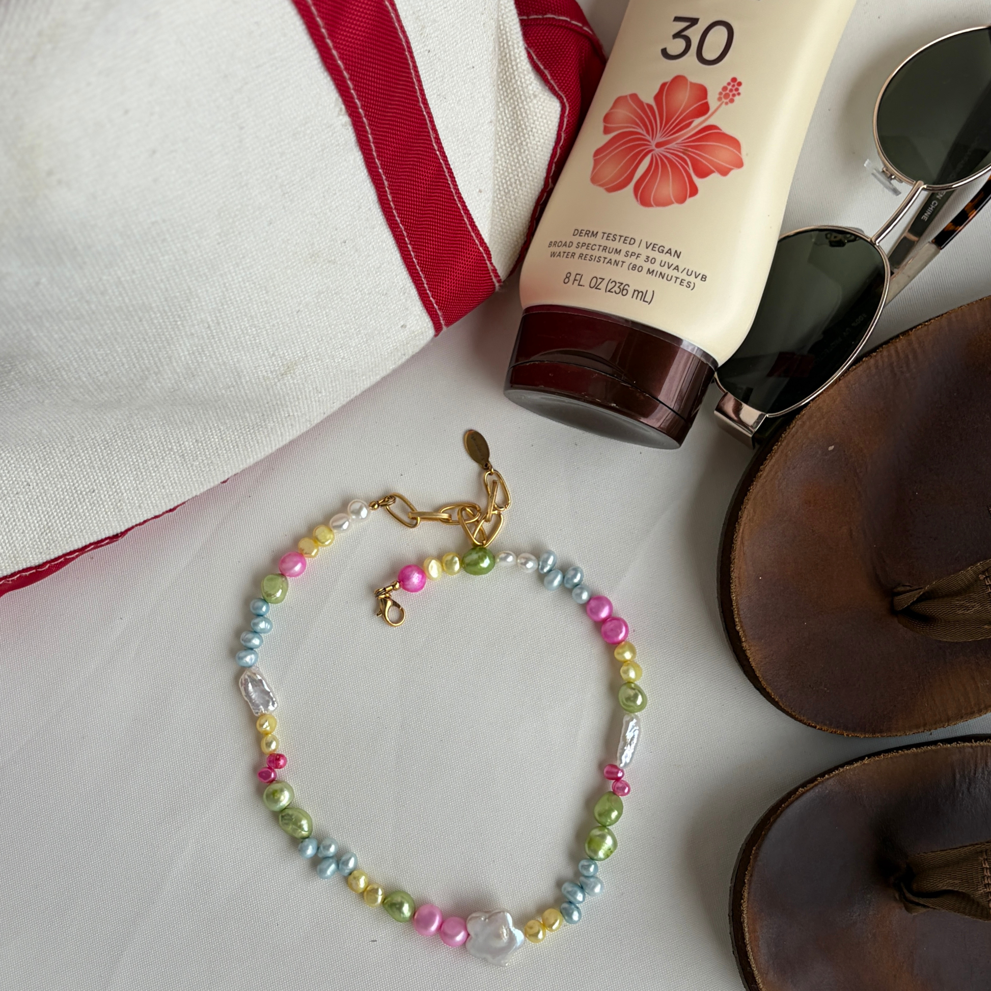 Colorful pearl bracelet beside a red-trimmed bag, SPF 30 sunscreen with a hibiscus, sunglasses, and brown sandals, evoking a relaxed beach vibe.