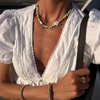 A person wearing a white, textured v-neck blouse and layered, colorful pearl necklaces. The mood is stylish and summery, accented by a gold bracelet and ring.