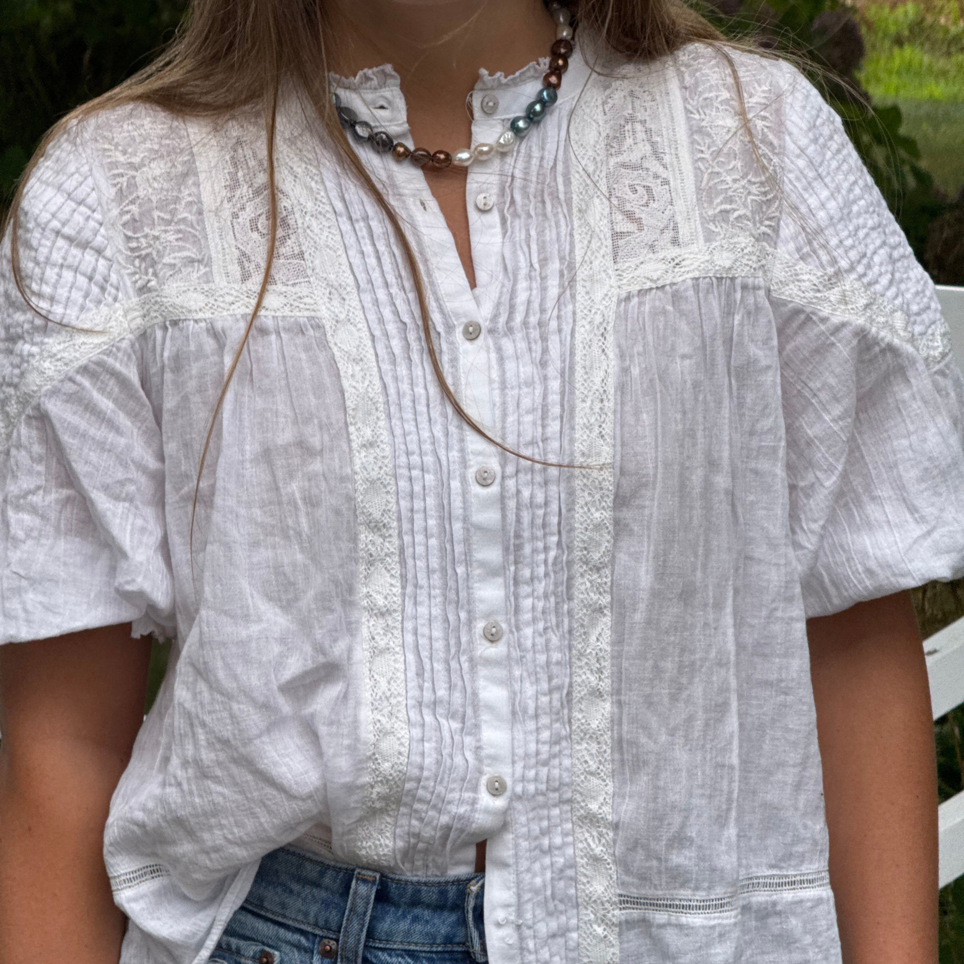 A person wearing a white short-sleeve blouse with lace trim, blue jeans in an outdoor setting. With a brown, blue, and white pearl necklace.