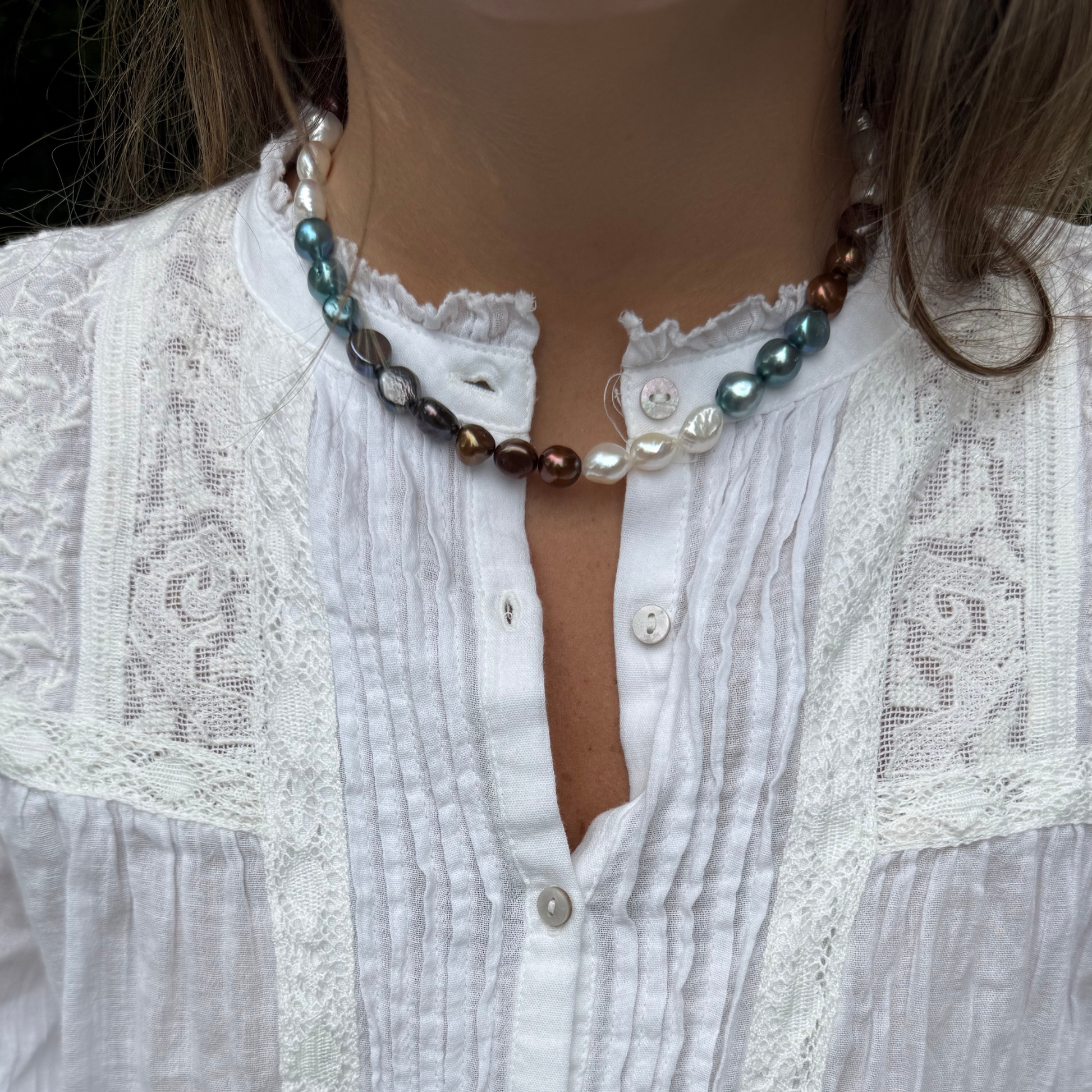A close-up of a person wearing a pearl necklace with white, teal, and brown over a white lace blouse.