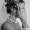 Black and white image of a woman with an elegant hairstyle, wearing pearl earrings, necklace, and bracelet. Her hand gently touches her face, exuding grace.
